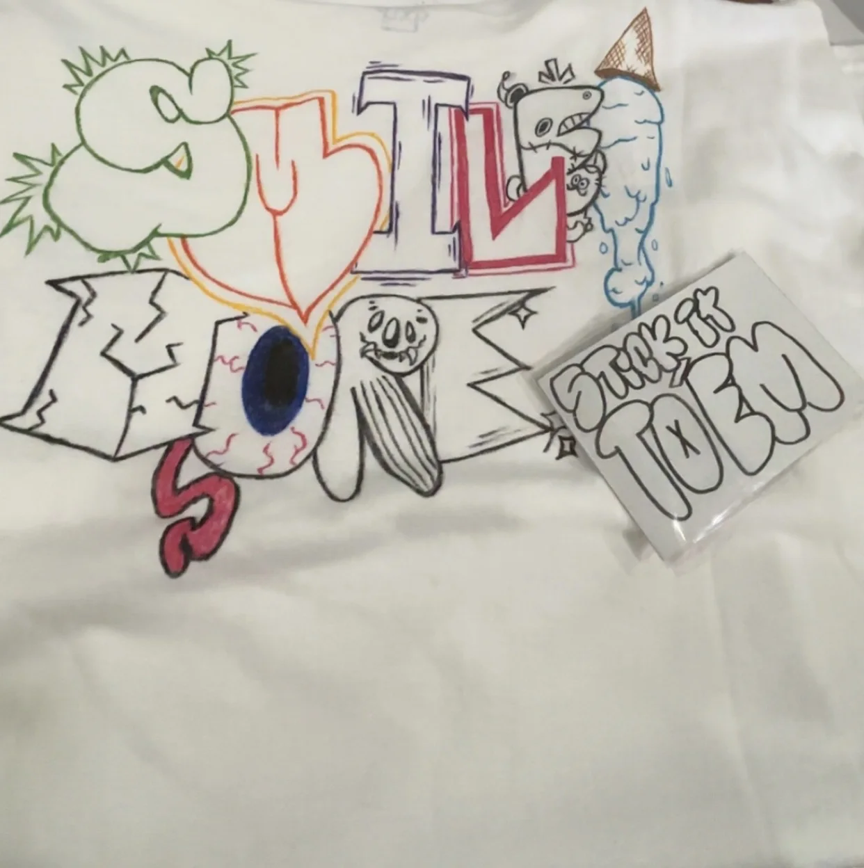 White shirt with a crowned smiling face, X eye, and spiral eye